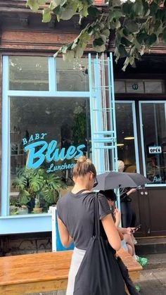 a woman holding an umbrella walking past a blue store front with people sitting on benches