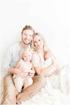 a man and woman holding a baby while sitting on top of a white bed next to each other