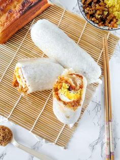 two burritos sitting on top of a bamboo mat next to bowls of food