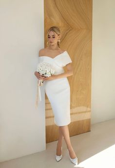 a woman in a white dress is holding a bouquet and standing against a wall with wood paneling