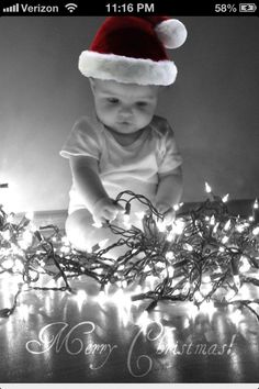 a black and white photo of a baby wearing a santa hat sitting in front of christmas lights