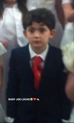 a young boy in a suit and tie standing next to other people wearing white dresses