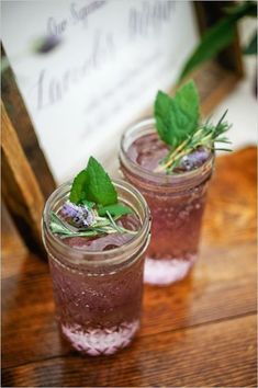 two mason jars filled with purple liquid and topped with fresh herbs, sitting on a wooden table