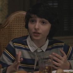 a boy sitting at a table with food in front of him and talking to someone