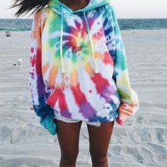 a woman standing on top of a sandy beach wearing a tie dye hoodie and shorts