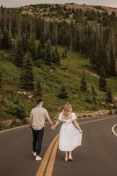 a man and woman holding hands walking down the road in front of some pine trees