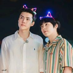 two young men standing next to each other in front of a dark background with colorful lights on their head