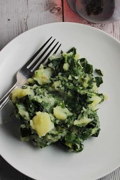 a white plate topped with greens and potatoes next to a glass of wine on top of a wooden table