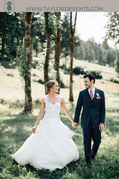 a bride and groom holding hands walking through the grass in front of some tall trees