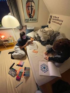 two people sitting on a bed in a room with lots of clutter and art supplies