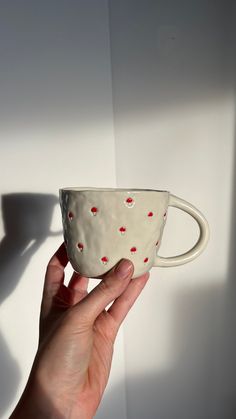 a hand holding a white cup with red dots on it in front of a wall