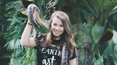 a woman holding a snake in her right hand while standing next to trees and plants