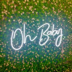 the word oh baby written in neon lights on a wall covered with green grass and white flowers