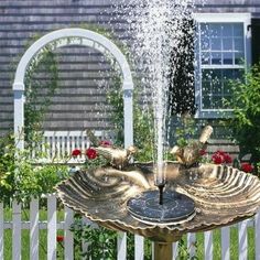 a bird bath fountain in front of a house with roses around it and two birds sitting on the bowl