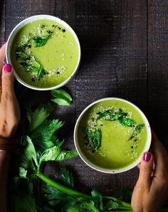 two hands holding bowls of green soup on top of a wooden table with parsley