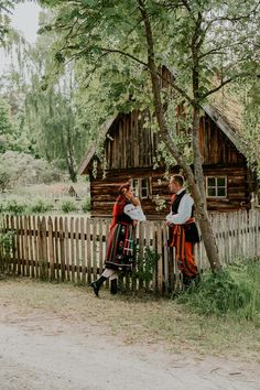 two people dressed in traditional clothing standing next to a wooden fence with a log cabin behind them