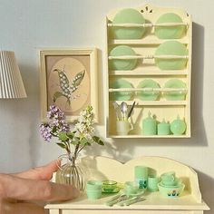 a hand is holding a vase with flowers in it next to a shelf filled with plates and cups