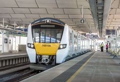 a yellow and white train pulling into a station with people standing on the platform next to it