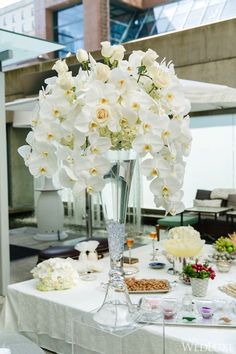 white flowers in a tall glass vase on top of a table with snacks and condiments