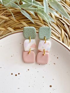 two pink and green square earrings sitting on top of a white plate next to a plant