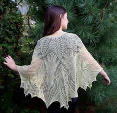 a woman wearing a white knitted shawl standing in front of some pine trees