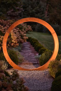 a large circular sculpture sitting in the middle of a garden next to a path and bushes