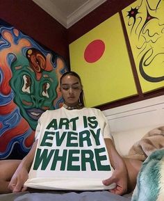 a woman sitting on her bed holding up a t - shirt that says art is everywhere