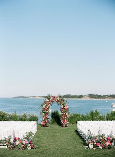 an outdoor ceremony setup with white chairs and colorful flowers on the grass near the water