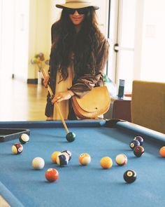 a woman standing next to a pool table holding a cue and wearing a fedora