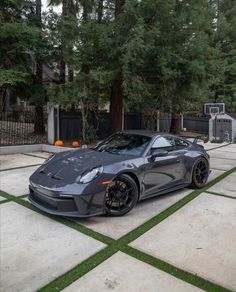 a gray sports car parked on top of a driveway next to a basketball hoop and trees