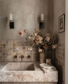 a bathroom sink with flowers in a vase on the counter next to two wall sconces