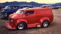 an old red truck parked in a parking lot next to other cars and trucks with mountains in the background