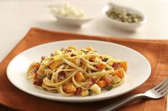 a white plate topped with pasta and bacon on top of a brown napkin next to a fork