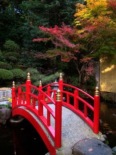 a red bridge over a small stream in a park with trees and rocks around it