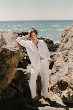 a man standing on rocks near the ocean with his hands behind his head and looking at the camera