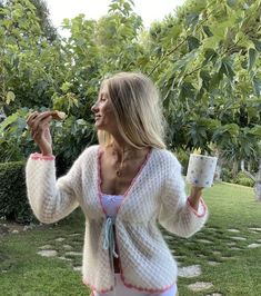 a woman in a white cardigan eating food and drinking from a cup on the grass