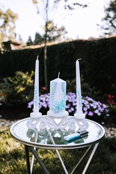 two white candles sitting on top of a table next to purple flowers and a blue pen