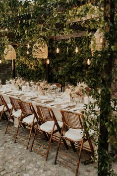 a long table set up with white linens and greenery for an outdoor dinner