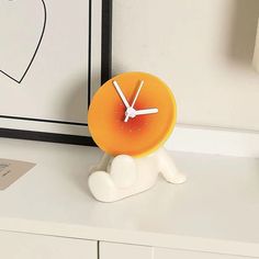 an orange clock sitting on top of a white desk next to a black framed heart