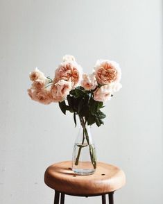 some pink flowers are in a clear vase on a small wooden table next to a white wall