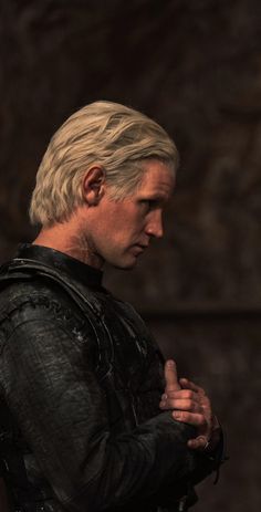 the white haired man is standing in front of a stone wall with his hands clasped