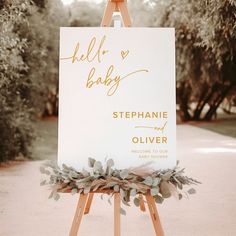 an easel with a sign that says hello baby on it and greenery around the edge