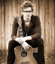 a young man sitting on the ground with his guitar in hand and looking at the camera