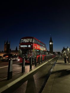 a red double decker bus driving down a street next to a tall clock tower at night