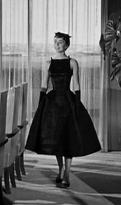 a woman in a black dress and hat walking down a hall with chairs around her