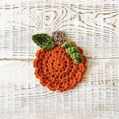 an orange crocheted brooch sits on a white wooden surface with green leaves