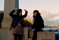 two women standing next to each other in front of a tall stone pillar with a city skyline behind them