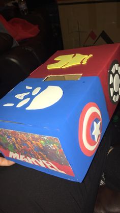 a child's hand is holding up a cardboard box with captain america on it