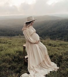 a pregnant woman wearing a long dress and hat sitting on a chair in a field