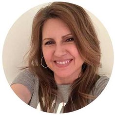 a woman with long brown hair wearing a gray shirt and hoop earrings smiles at the camera